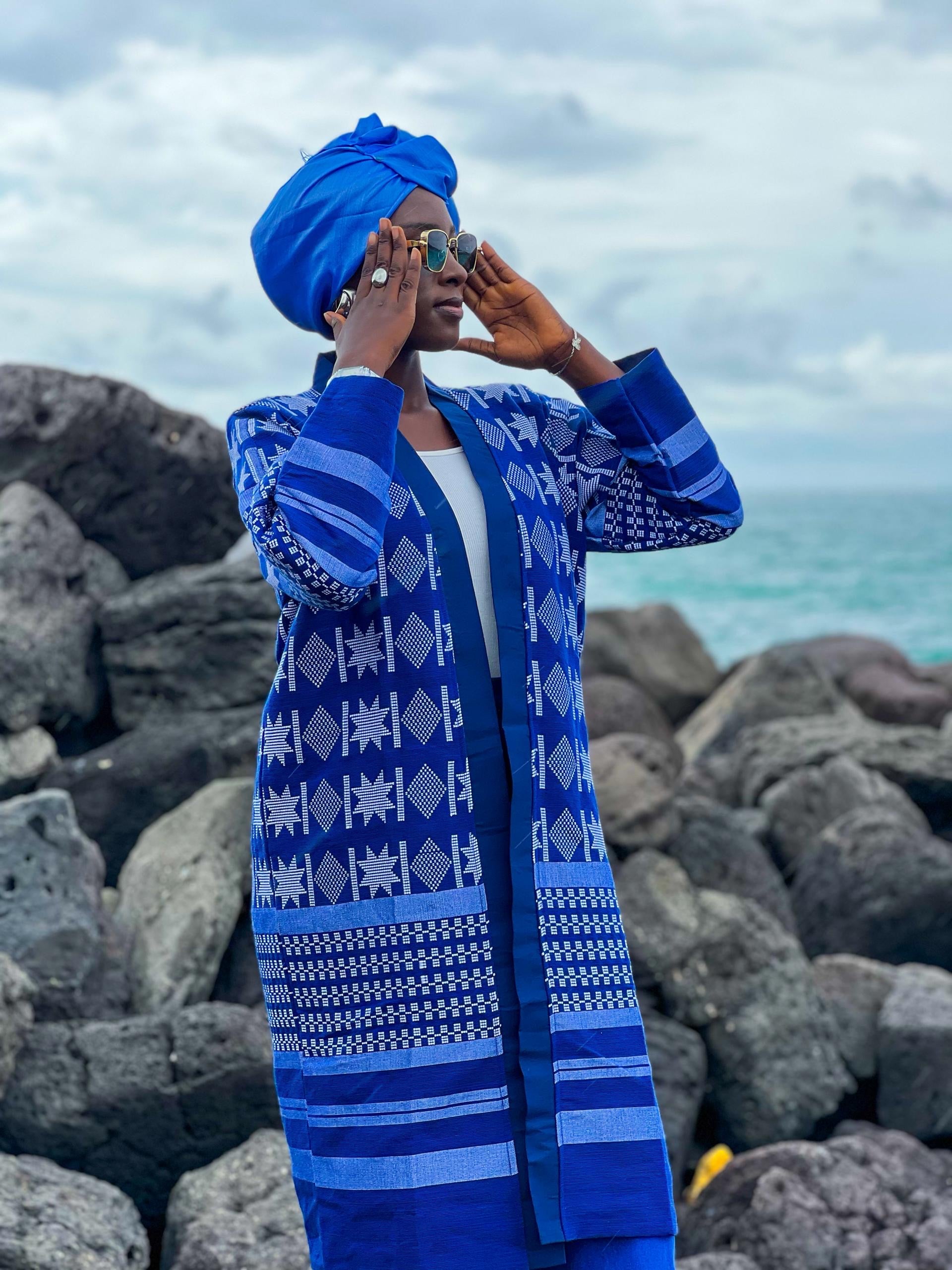 Woman wearing a Kanoo Hand Woven Jacket and blue headwrap, standing by rocks near the ocean.