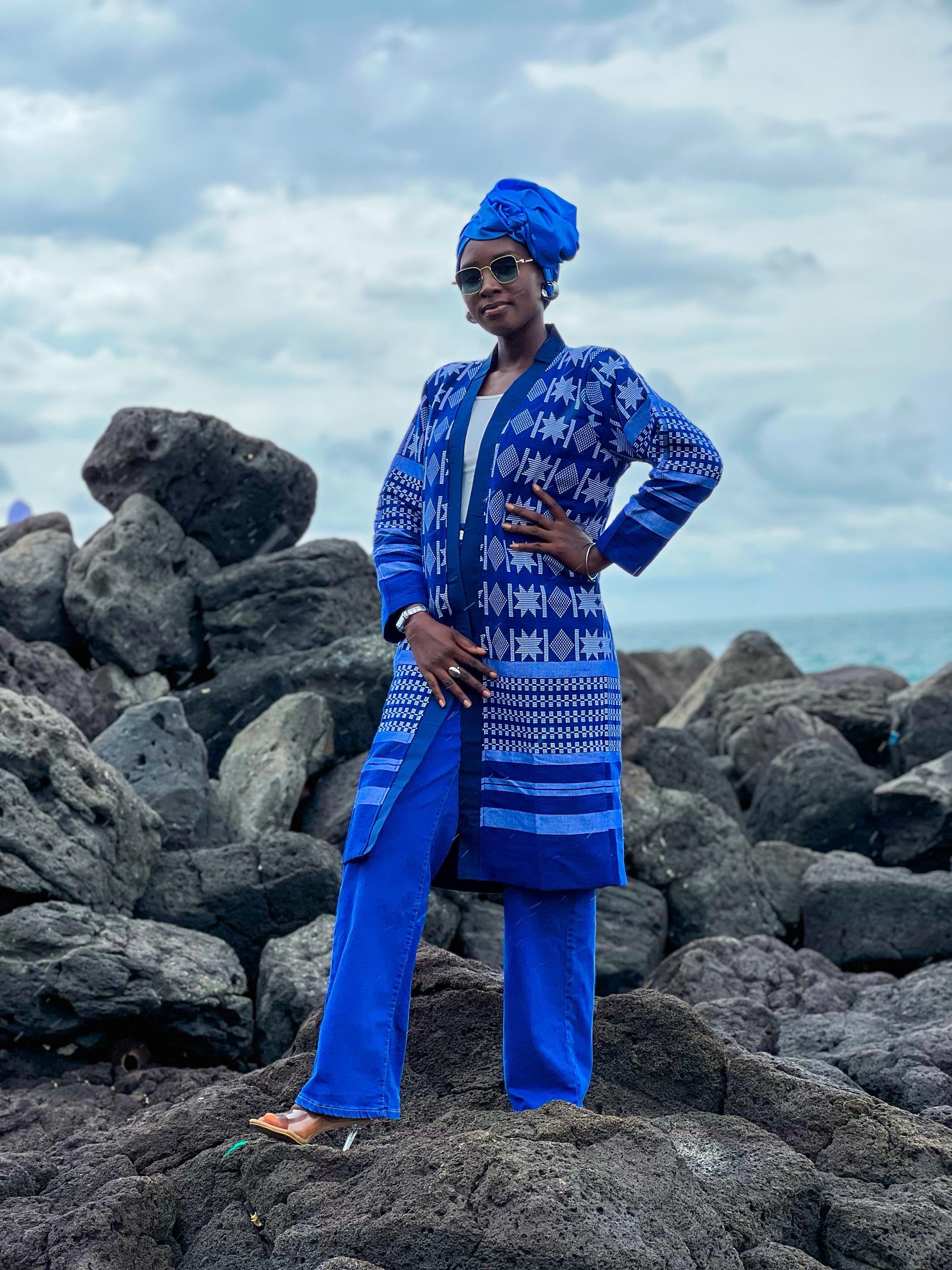 Woman wearing Kanoo Hand Woven Jacket on rocky beach, showcasing vibrant blue geometric patterns and cultural heritage.