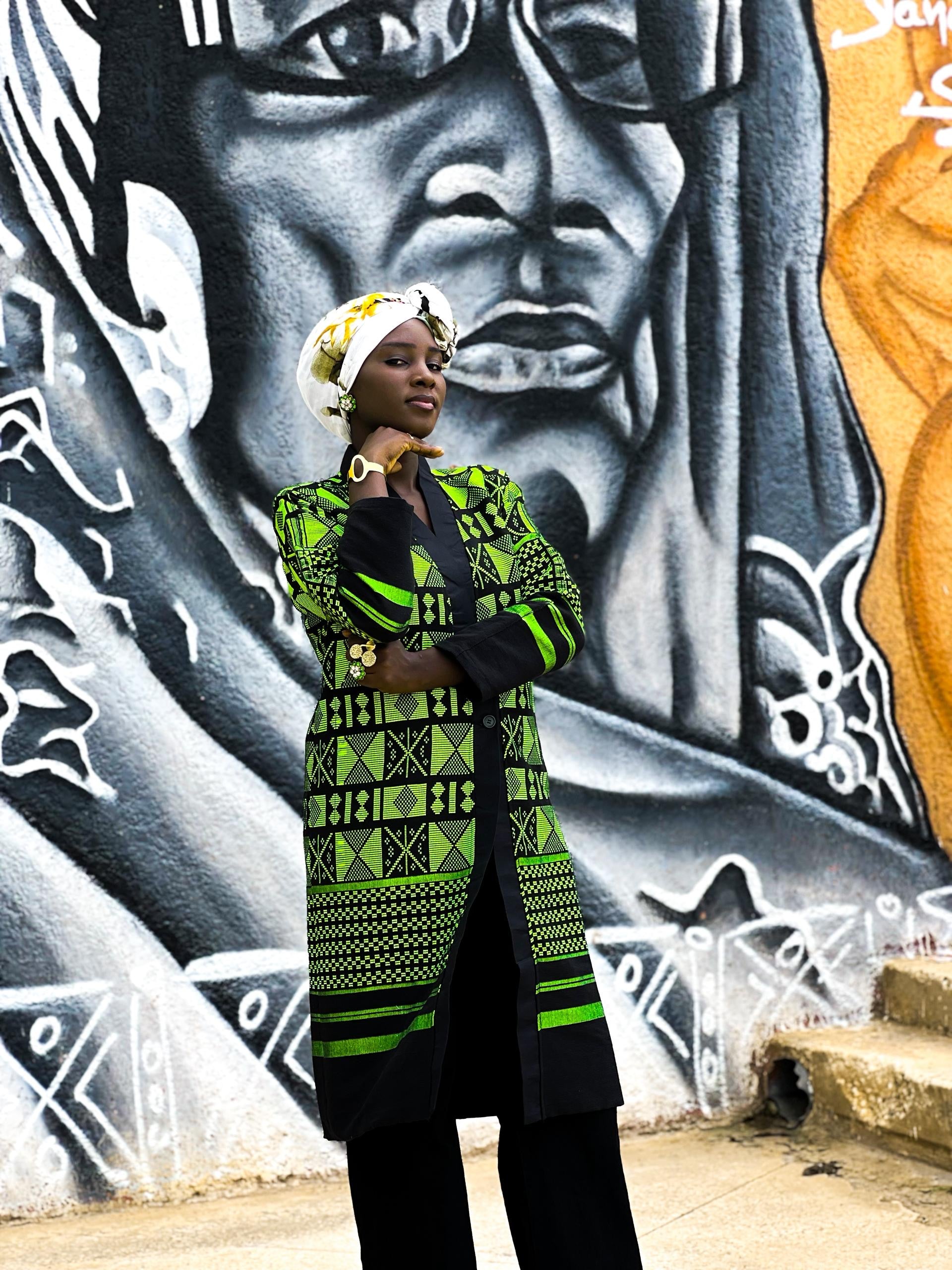 Kanoo Hand Woven Jacket model poses in front of a vibrant mural, showcasing Senegalese craftsmanship and cultural heritage.